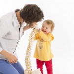 Mom physiotherapist with laughing baby, showing her a model of a spine