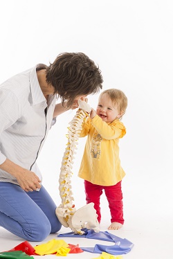 Mom physiotherapist with laughing baby, showing her a model of a spine
